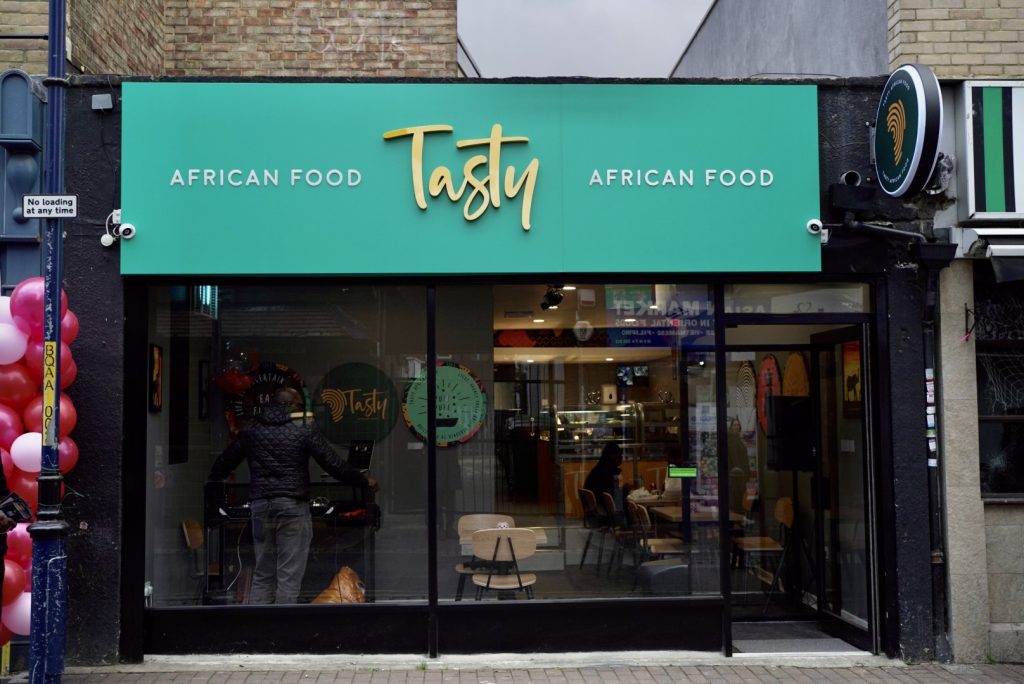 The image shows the storefront of Tasty African Food a restaurant with a green awning large windows and a visible interior with seating