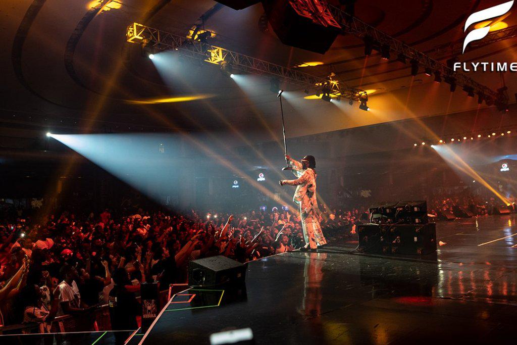 A performer on stage singing to a large audience under bright stage lights with an event banner in the background